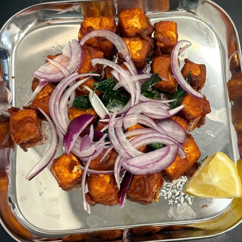 Crispy fried paneer cubes with red onion slices, curry leaves, and a lemon wedge on a silver tray.