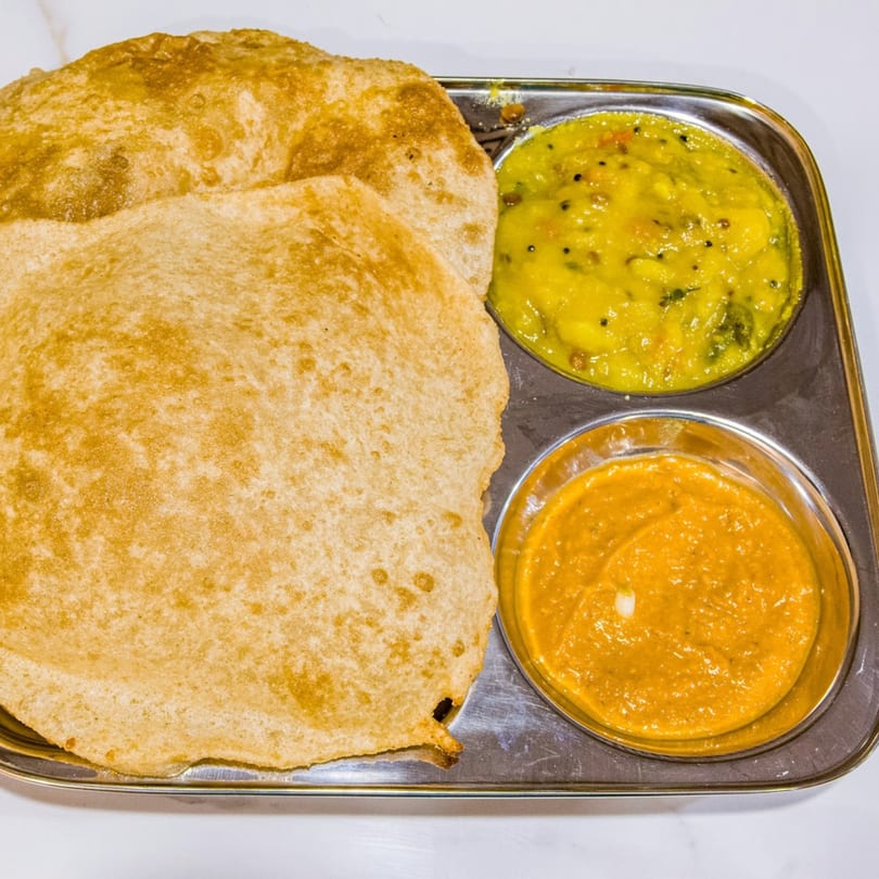 Two golden-brown puri breads on a metal tray with yellow potato curry and orange chutney.