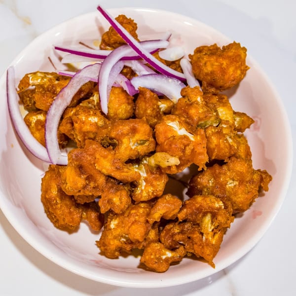 Crispy fried cauliflower florets, golden brown and seasoned, topped with thinly sliced red onions in a white bowl.