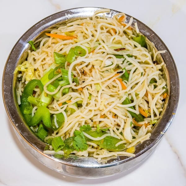 A close-up of a metal bowl filled with vegetable chow mein noodles, green peppers, and carrots.