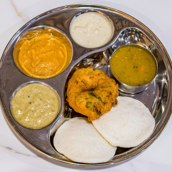 A stainless steel thali with idli, vada, sambar, and three chutneys on a white marble surface.