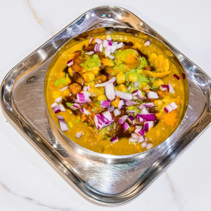 A bowl of Indian chaat with chickpeas, sauces, and chopped red onions on a silver tray.